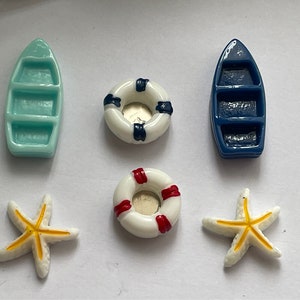 Op de afbeelding: Een verzameling miniatuurfiguren met een nautisch thema. De set bevat twee kleine boten, twee reddingsboeien en twee zeesterren. De boten zijn zeegroen en marineblauw. De reddingsboeien zijn wit met rode en marineblauwe accenten. De zeesterren zijn wit met gele details.