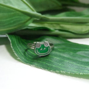 May include: A silver ring with a green enamel frog design. The ring is on a green leaf background.
