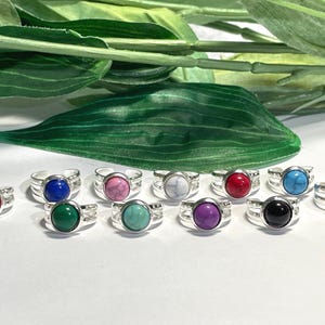 May include: A collection of twelve adjustable silver-tone rings, each featuring a round, smooth, marbled-look stone in a variety of vibrant colors including pink, blue, green, red, white, purple, and black, arranged in two rows on a white surface.