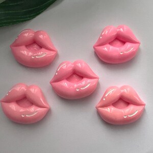 May include: Five pink lip-shaped embellishments with glossy highlights. The lips are a vibrant pink colour with a subtle sheen, perfect for crafting or decorative projects. The image is taken from a top-down perspective.