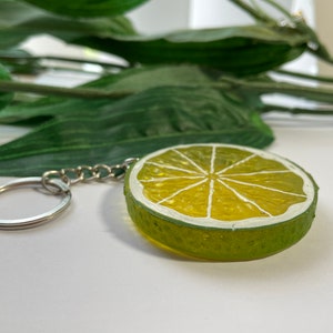 May include: A lime slice keychain with a silver chain and ring. The keychain features a green rind and a yellow interior with white segments. The design is reminiscent of a fresh lime, perfect for adding a touch of summer to your keys.