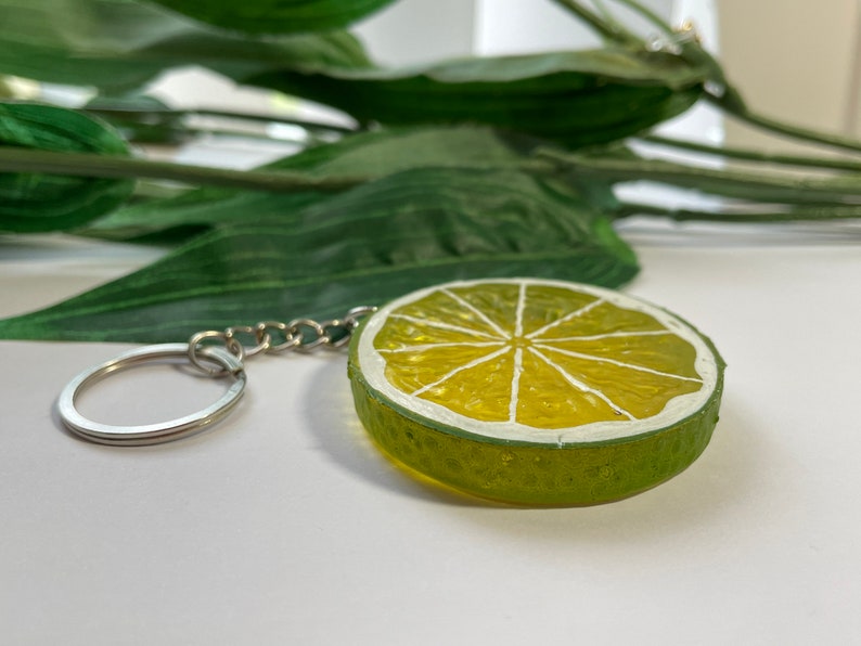 May include: A lime slice keychain with a silver chain and ring. The keychain features a green rind and a yellow interior with white segments. The design is reminiscent of a fresh lime, perfect for adding a touch of summer to your keys.