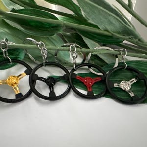 Può includere: Quattro portachiavi a forma di volante nero con centri di colori diversi. I centri sono dorati, rossi, neri e argentati.