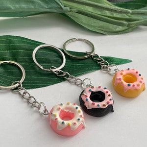 Può includere: Tre portachiavi argentati con ciondoli a forma di ciambella. Le ciambelle sono rosa, nere e gialle con glassa bianca e codette colorate.