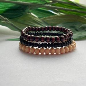 May include: Three stacked wooden bead bracelets. The top bracelet is brown, the middle bracelet is black, and the bottom bracelet is natural wood.