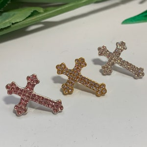 May include: Three rhinestone cross charms in silver, gold, and pink. The crosses are all the same size and shape, and they are decorated with small rhinestones.