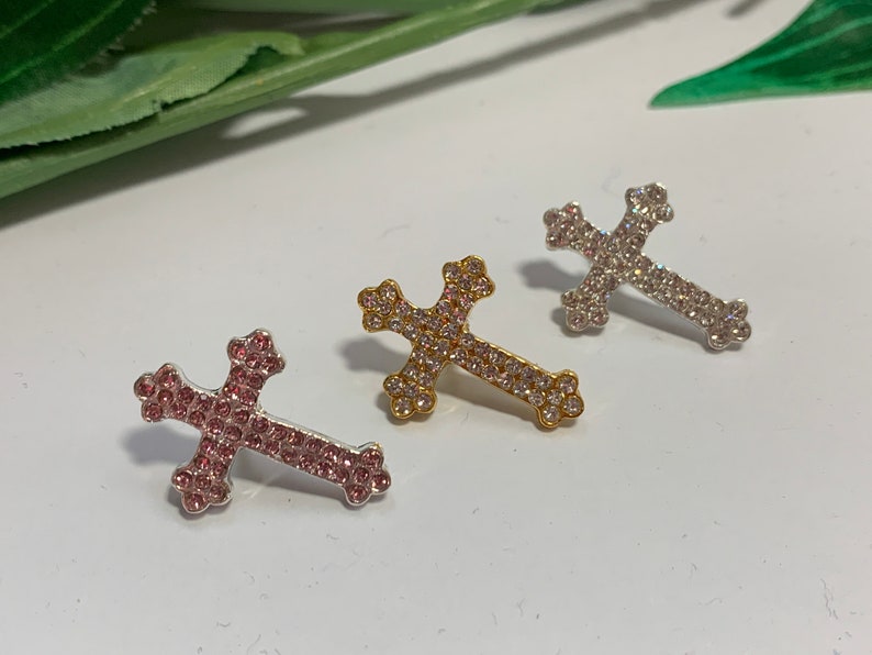 May include: Three rhinestone cross charms in silver, gold, and pink. The crosses are all the same size and shape, and they are decorated with small rhinestones.