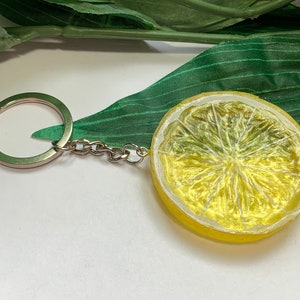 May include: A lemon slice keychain with a silver keyring and chain. The lemon is yellow with a textured surface, and the interior is a lighter yellow with visible segments. A green leaf is in the background.