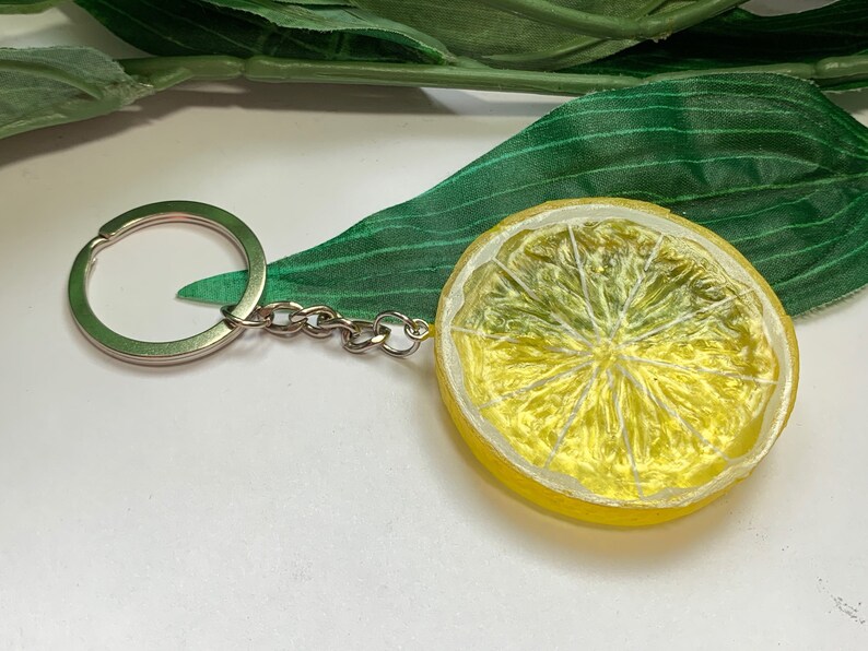 May include: A lemon slice keychain with a silver keyring and chain. The lemon is yellow with a textured surface, and the interior is a lighter yellow with visible segments. A green leaf is in the background.