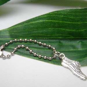 May include: A silver charm depicting a running shoe, attached to a ball chain. The charm shows detailed features of a trainer. The chain is made of small, connected silver balls and is attached to a clasp.