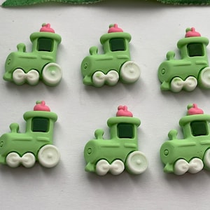 May include: Six green train-shaped embellishments with white wheels and a pink top hat. The train has a dark green window and a small chimney. These decorative pieces are suitable for crafts and scrapbooking.