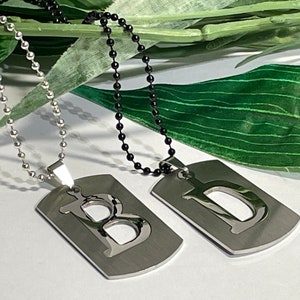 May include: Two silver dog tag necklaces with the letters "B" and "D" cut out of the centre. The necklaces are on a white surface with green leaves behind them. One necklace has a silver chain and the other has a black chain.