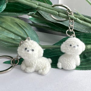 May include: Two white fluffy dog keychain charms with black eyes. The charms are attached to silver key rings.