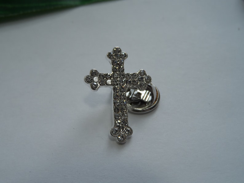May include: A silver cross pin with clear rhinestones. The pin has a safety clasp on the back.