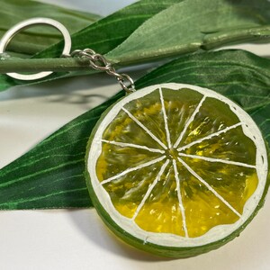 May include: A lime slice keychain with a silver keyring and chain. The lime slice is painted with green and yellow hues, and white accents. The keychain is set against a backdrop of green leaves.