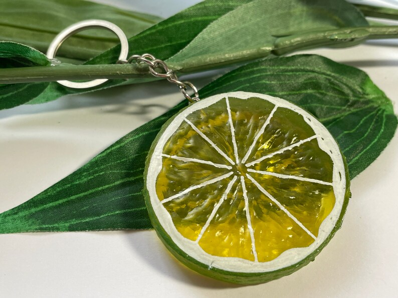 May include: A lime slice keychain with a silver keyring and chain. The lime slice is painted with green and yellow hues, and white accents. The keychain is set against a backdrop of green leaves.