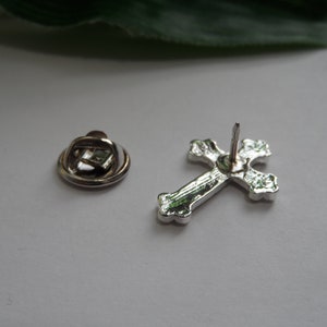 May include: Silver cross pin with a decorative design. The pin has a safety clasp on the back.