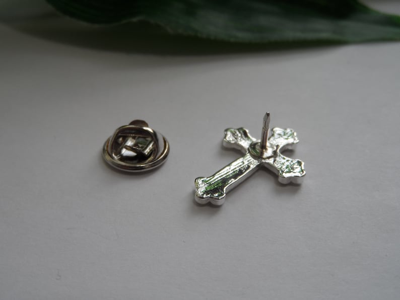 May include: Silver cross pin with a decorative design. The pin has a safety clasp on the back.