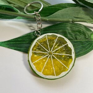 May include: A lime slice keychain with a silver chain and ring. The lime slice is painted with yellow and white details, and a green rind. The keychain is a fun accessory.