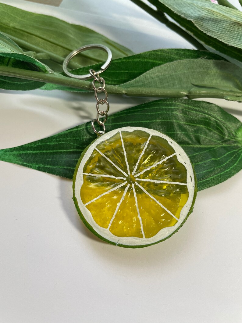 May include: A lime slice keychain with a silver chain and ring. The lime slice is painted with yellow and white details, and a green rind. The keychain is a fun accessory.