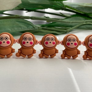 May include: Five brown monkey-shaped charms with tan faces, pink cheeks, and large black eyes. Each charm has a small, curled tail. The charms are arranged in a row on a white surface.
