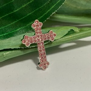 May include: A silver cross charm with pink rhinestones. The cross is a religious symbol and is often used in jewelry.