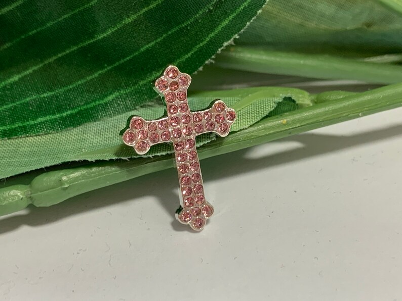 May include: A silver cross charm with pink rhinestones. The cross is a religious symbol and is often used in jewelry.
