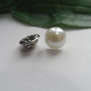 May include: A round white pearl button with a smooth, lustrous finish. Next to it lies a silver-toned metal button back featuring a textured, studded edge and a central loop for attachment, showcasing the construction of the fastener.