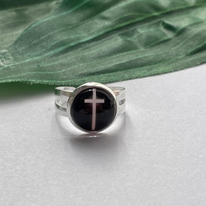 Könnte beinhalten: Ein silberner Ring mit einem schwarzen und weißen Kreuzdesign. Der Ring hat einen runden, schwarzen Cabochon mit einem weißen Kreuz in der Mitte.
