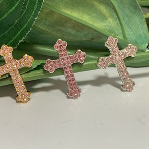 May include: Three cross charms with pink rhinestones. The crosses are gold, silver, and silver with a pink hue.