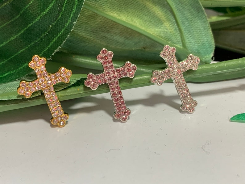 May include: Three cross charms with pink rhinestones. The crosses are gold, silver, and silver with a pink hue.