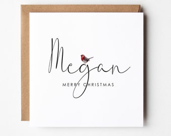 Op de afbeelding: Een witte kerstkaart met de naam "Megan" geschreven in zwarte cursieve letters. Een kleine roodborst zit boven de naam. De tekst "Prettige Kerst" staat in zwart onder de naam.