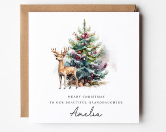 Op de afbeelding: Een kerstkaart met een aquarel-illustratie van een hert dat naast een gedecoreerde kerstboom staat. De kaart luidt "Prettige Kerstmis aan onze mooie kleindochter Amelia."