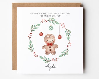 Op de afbeelding: Een witte kerstkaart met een aquarel-illustratie van een peperkoekman in een groene krans met rode bessen en groene en rode ornamenten. De tekst "Merry Christmas to a special granddaughter" staat bovenaan de kaart en de naam "Ayla" staat onderaan.