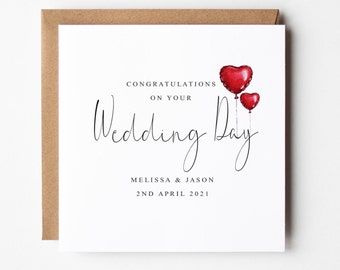 May include: A white card with the text "Congratulations on your Wedding Day" in black script. Below the text is "Melissa & Jason" and "2nd April 2021". Two red heart-shaped balloons are floating above the text.