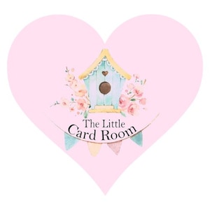 May include: A pink heart-shaped sign with a light blue birdhouse and floral accents. The sign reads "The Little Card Room".