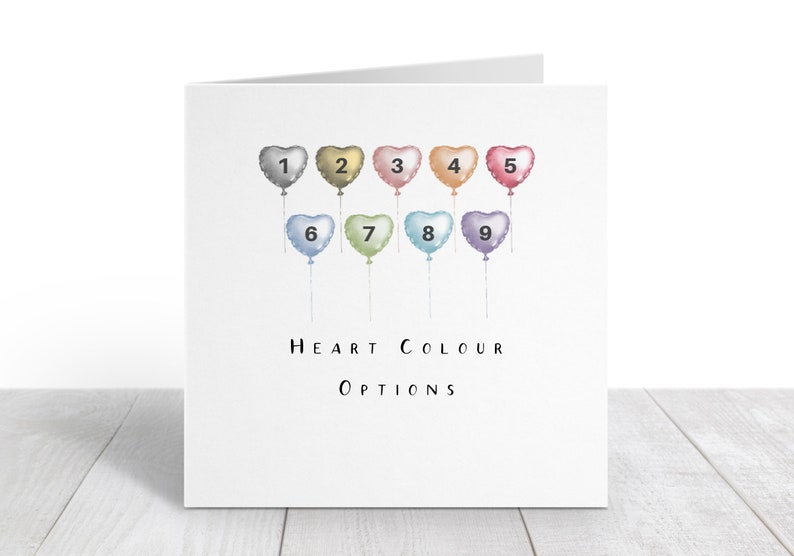 K&ouml;nnte beinhalten: Eine Karte mit neun herzf&ouml;rmigen Ballons in verschiedenen Farben, nummeriert von 1 bis 9. Der Text "HEART COLOUR OPTIONS" ist unter den Ballons gedruckt.