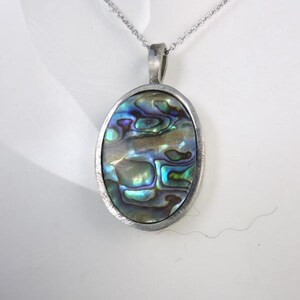 Könnte beinhalten: Ovale Anhänger Halskette mit einem silberfarbenen Metallrahmen. Der Anhänger zeigt eine schillernde Abalone-Schale mit Blau-, Grün- und Lilatönen. Die Halskette hat eine zarte Silberkette.