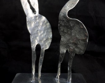 Op de afbeelding: Een zilveren metalen sculptuur van twee vogels die op een rechthoekige basis staan. De vogels zijn gestileerd en hebben een gehamerde textuur.