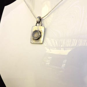 Puede incluir: Un collar de plata con un colgante cuadrado que presenta una piedra redonda gris en el centro.