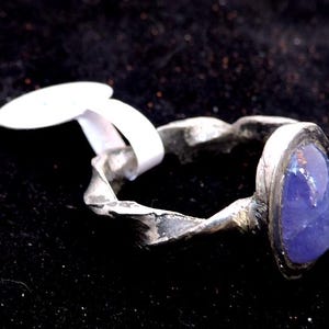 Peut inclure: Une bague en argent avec une grande pierre précieuse ovale de couleur violette sertie dans la bande. La bague a une finition texturée et martelée.