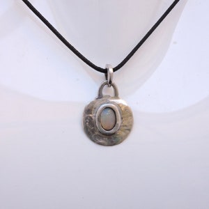 Peut inclure: Un collier avec un pendentif en argent rond et martelé, orné d'une opale blanche. Le collier est sur un cordon noir.