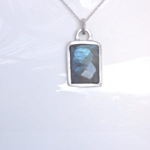 Puede incluir: Un collar plateado con un colgante rectangular con una piedra iridiscente azul y gris brillante. El colgante está engastado en un marco plateado y cuelga de una fina cadena. La joya se muestra sobre un fondo blanco.