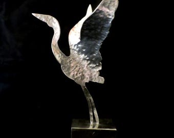 Op de afbeelding: Een zilveren metalen sculptuur van een kraanvogel met uitgespreide vleugels, staande op een vierkante basis.