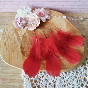 Peut inclure: Cinq plumes rouges sur une surface en bois avec une dentelle blanche en premier plan. Les plumes sont disposées en éventail. Il y a aussi des fleurs en feutre rose et blanc sur la surface.