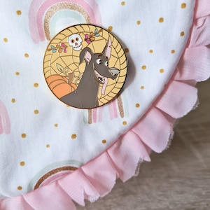 May include: A gold-colored enamel pin featuring a cartoon dog with a skull and crossbones on a yellow background. The dog is wearing a floral garland and is surrounded by a web.