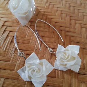 May include: A set of white origami rose earrings and a ring. The earrings are made of white paper and have silver hooks. The ring is also made of white paper and has a silver band.