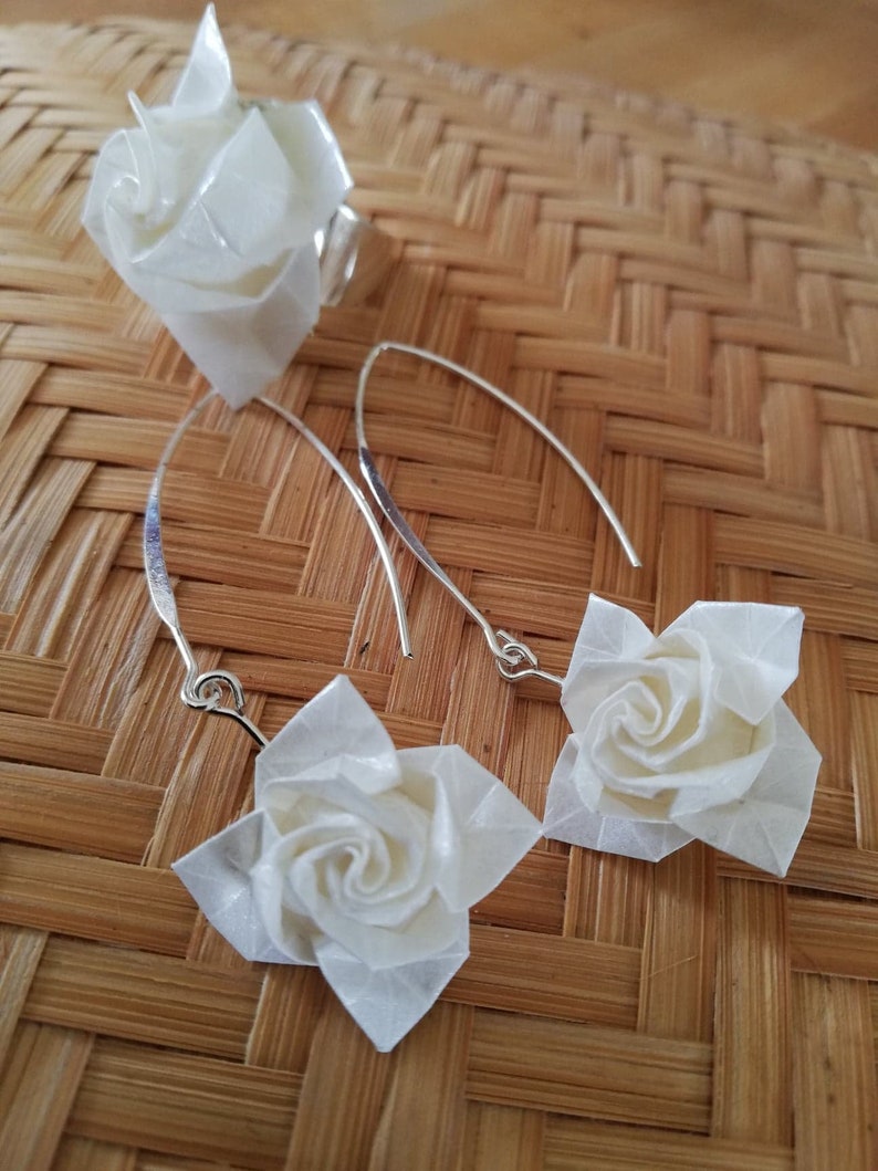 May include: A set of white origami rose earrings and a ring. The earrings are made of white paper and have silver hooks. The ring is also made of white paper and has a silver band.
