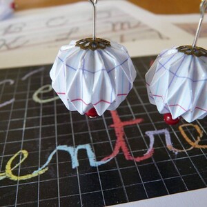 Op de afbeelding: Een paar oorbellen met witte papier origami bollen met rode kralen en een zilveren metalen accent. De bollen hebben een blauw en rood rasterpatroon.