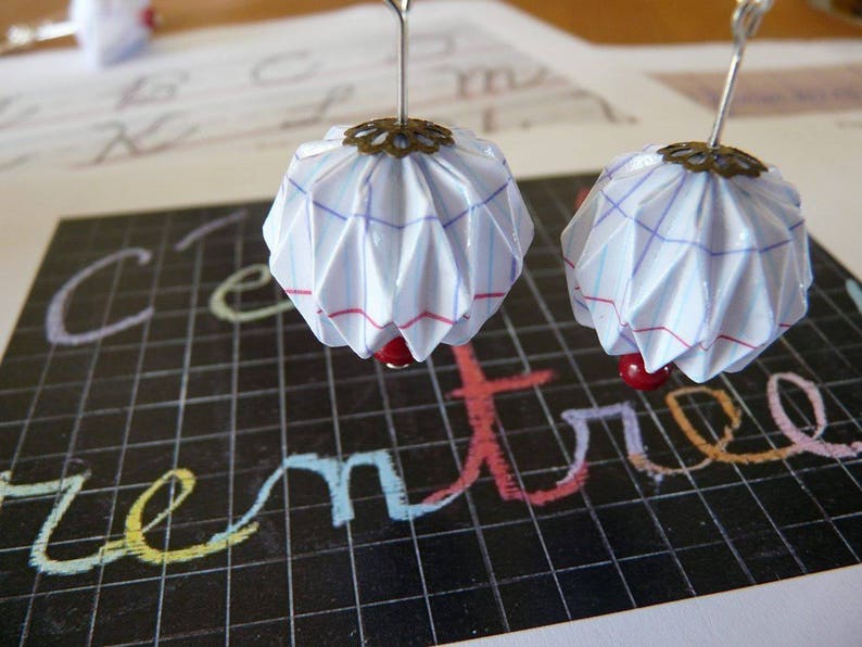 Op de afbeelding: Een paar oorbellen met witte papier origami bollen met rode kralen en een zilveren metalen accent. De bollen hebben een blauw en rood rasterpatroon.
